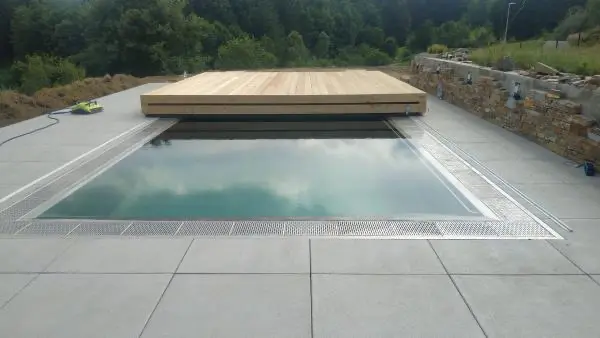 <p>Zde je vidět příkladná ukázka toho, jak designové zastřešení Slide Wood spojuje celý koncept zahrady a rodinného domu. Zastřešení je tvořeno ze dvou dílů, přičemž menší díl zajíždí do většího dílu a oba díly se odsunou za hranu bazénu. Na povrch zastřešení se použily přírodní dřevěná terasová prkna, která se ukazují taktéž na fasádě domu. Přirozenou přírodní atmosféru podtrhává kámen kolem terasy u bazénu.</p>
